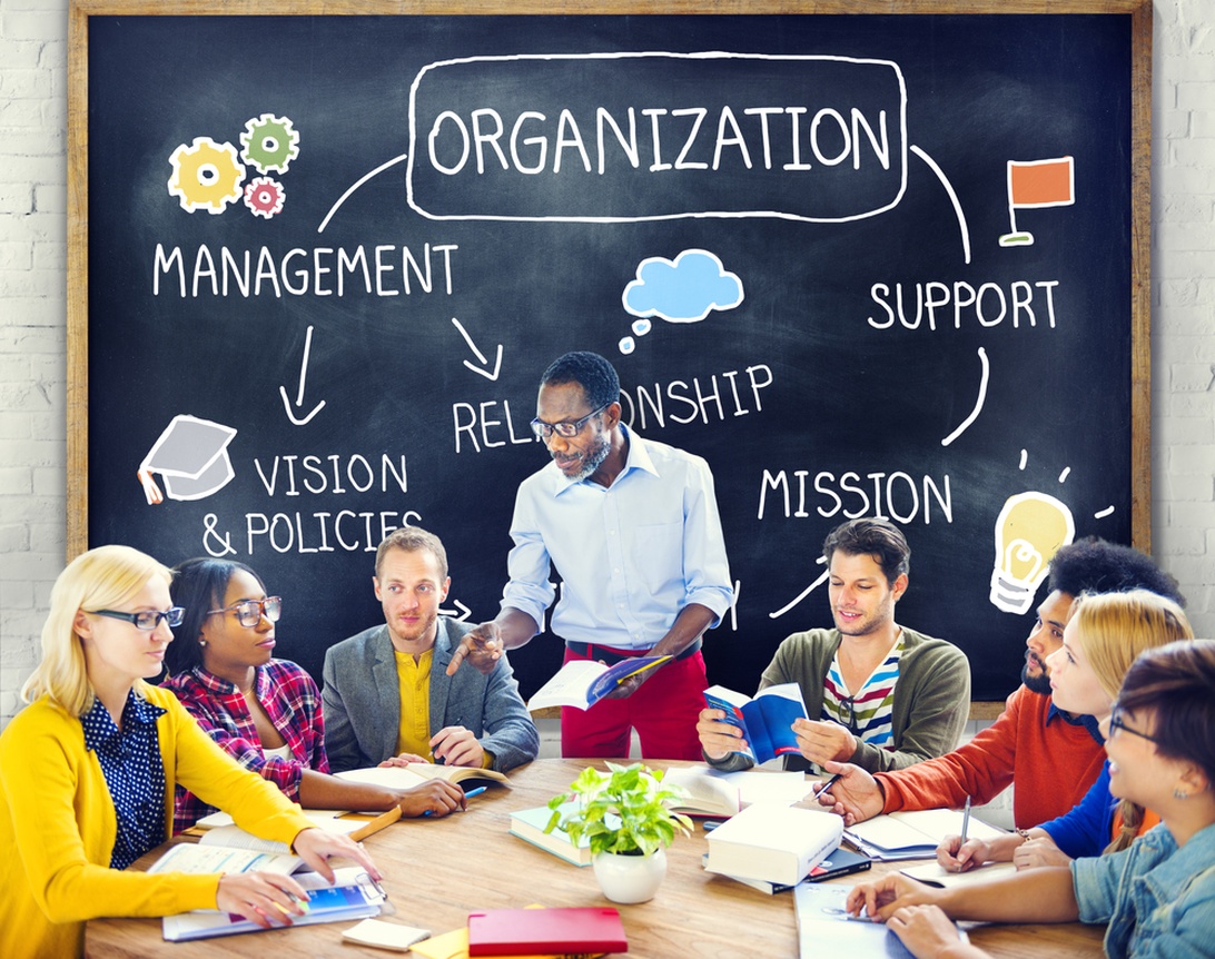 A diverse team collaborates on strategy, with concepts like 'VISION' and 'MISSION' on a blackboard behind them.